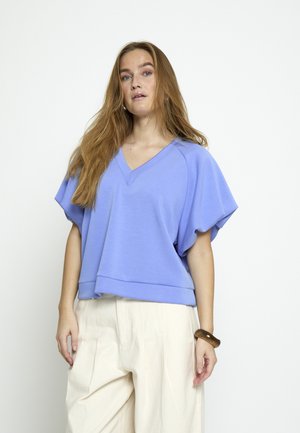 Kvinde med langt lyst brunt hår iført en løs lyseblå V-hals bluse og cremefarvede bukser med vide ben, stående mod en hvid baggrund.