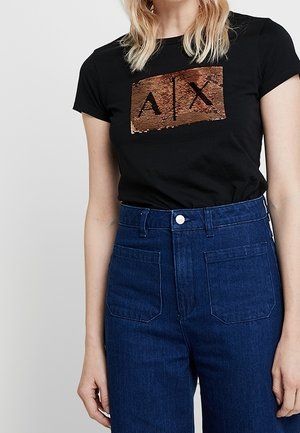 Kvinde iført en sort T-shirt med et kobberfarvet palietlogo A|X og mørkeblå højtaljede jeans, stående mod en hvid baggrund.