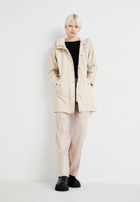 Veste imperméable beige avec capuche, fermeture éclair et boutons-pression, poches latérales ; associée à un pantalon beige clair et des chaussures à plateforme noires.