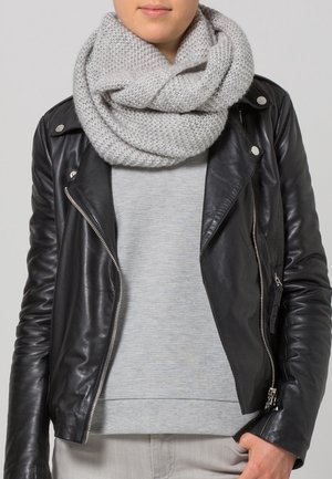 Personne portant une écharpe infinie en maille grise, une veste en cuir noire, un pull gris et un pantalon clair, main partiellement dans la poche.