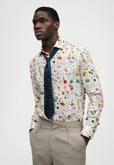 Virágmintás ing fehér alapon, színes virágokkal és rovarokkal, navy kék nyakkendővel, amelyen apró minták találhatók, és világos bézs nadrággal.