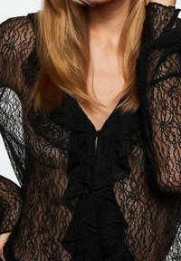 Blouse noire en dentelle transparente avec détail à volants sur le devant, portée par une personne aux longs cheveux blonds, main près de la tête.