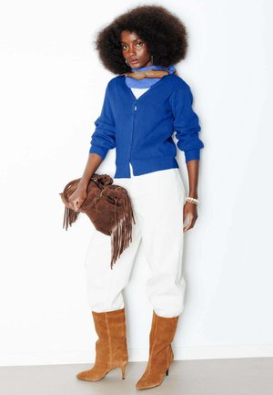 Femme avec une coiffure afro portant un cardigan bleu, une écharpe multicolore, un pantalon blanc, des bottes en daim camel, tenant un sac marron à franges sur fond blanc.