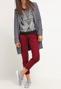 Grå lång cardigan över en svart- och vitmönstrad tröja, i kombination med bordeauxfärgade slim-fit-byxor och grå sportskor.