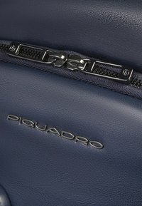 Marineblauwe leren tas met een textuurafwerking, voorzien van een zilverkleurige rits en een reliëf met het merk op de zijkant. Duurzaam en functioneel ontwerp.