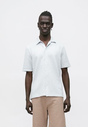 Svetlosivé tričko s krátkym rukávom z textúrovaného materiálu, s golierom a zapínaním na gombíky, doplnené o svetlo béžové plisované nohavice.