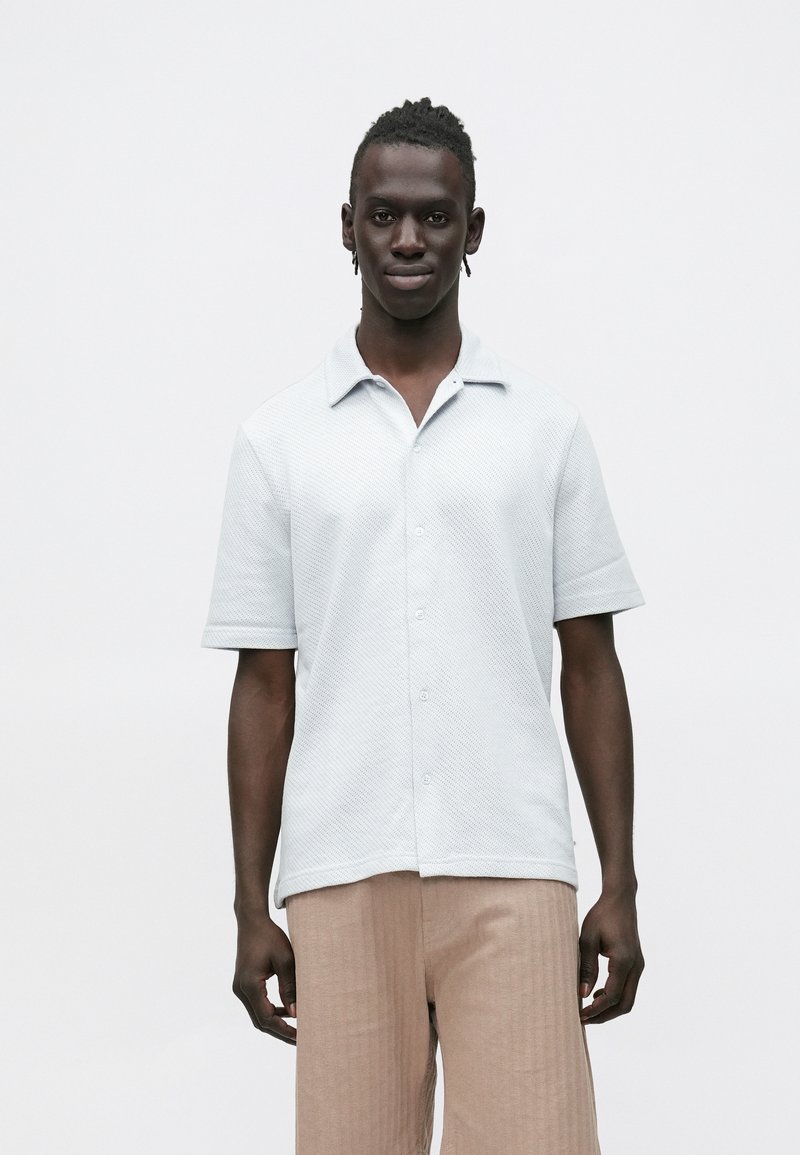 Lysegrå kortermet skjorte i teksturert stoff, med krage og knappelukking, kombinert med lysebeige bukser med folder.