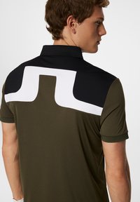 Miesten polo-paita, jossa on tummanoliivinen pohja, musta kaulus ja valkoinen abstrakti kuvio selässä. Sujuva, kevyt kangas ja lyhyet hihat.