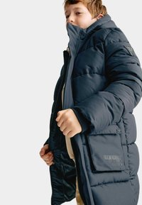 Marineblauwe gewatteerde jas met een hoge kraag, ritssluiting, geribde manchetten en een buitenzak met geprinte details. Zachte, gewatteerde structuur.