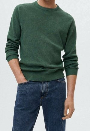 Mand iført grøn langærmet sweater og blå jeans med den ene hånd i lommen mod en ensartet lys baggrund.