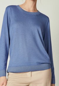 Pull en maille bleu clair avec col rond et poignets côtelés, coupe décontractée et texture douce. Associé à un pantalon beige.