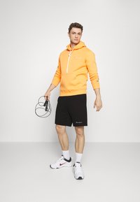Sudadera naranja con cierre de media cremallera y bolsillo frontal, combinada con pantalones cortos negros. Los accesorios incluyen una cuerda para saltar y zapatillas blancas.