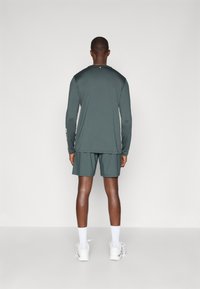 Σκούρο πράσινο αθλητικό μπλουζάκι με μακριά μανίκια και στρογγυλή λαιμόκοψη, σε συνδυασμό με ταιριαστά σορτς. Λευκά αθλητικά παπούτσια και κάλτσες ολοκληρώνουν το σύνολο.