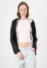 Jaqueta leve preta com capuz, combinada com uma blusa justa rosa claro com um logo brilhante "Hugo". Jeans de cintura alta azul claro.