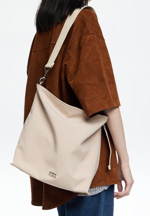 Beige slouchy tas van glad materiaal met een trekkoordsluiting en een verstelbare crèmekleurige schouderband; heeft een logo aan de onderkant.