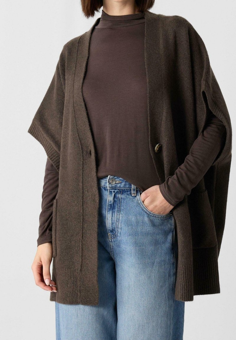 Femme portant un haut marron à manches longues sous un cardigan marron en maille ample avec poches, associé à un jean taille haute bleu clair, main dans la poche.