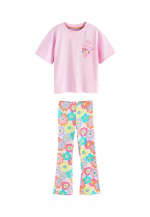 Copil cu păr lung purtând un tricou roz cu text colorat și pantaloni cu model floral, stând cu fața spre spate pe un fundal simplu.