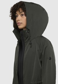 Giacca impermeabile verde scuro con cappuccio, dotata di zip completa, cordino regolabile e dettaglio del logo sulla manica.