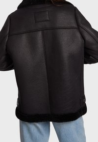 Svart läderjacka med en texturerad yta, som har en shearling-krage och en mjuk svart kant vid nederkant och ärmar.