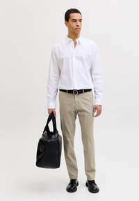 Mann iført hvit skjorte med knapper, beige bukser, svarte sko og belte, holder en svart Duffel-veske i høyre hånd, stående mot hvit bakgrunn.