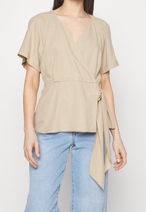 Frau trägt ein beiges Wickel-Oberteil mit kurzen Ärmeln, das in der Taille gebunden ist, kombiniert mit hellblauen Jeans vor einem einfarbigen Hintergrund.