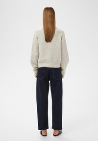 Osoba s rovnými dlouhými vlasy, oblečená ve světle šedém pleteném svetru, tmavě modrých širokých kalhotách a černých slip-on botách, otočená zády k bílé pozadí.