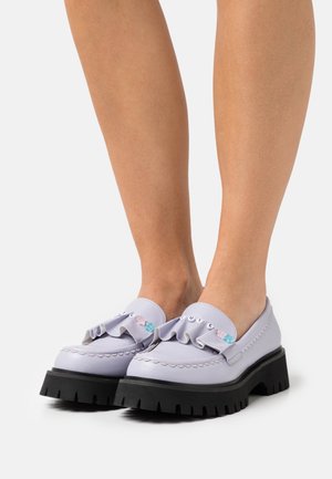 Mocassins lavande avec semelles épaisses noires portés pieds nus, décorés de volants et de perles formant le mot "MIGNON" avec des accents en forme d'étoile.