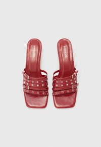 Mules rojos de punta abierta con múltiples diseños de tiras adornadas con tachuelas plateadas y hebillas. Textura suave con punta cuadrada y tacón bloque.