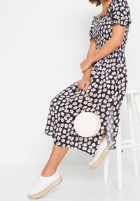 Frau sitzt auf einem weißen Hocker, trägt ein marineblaues, floral gemustertes Midikleid mit Puffärmeln, weiße Plateausneaker und hält eine runde, weiße, gesteppte Handtasche.