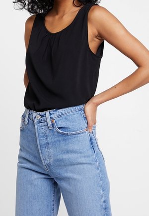 Kvinde iført en ærmeløs sort top stoppet ned i lyseblå jeans med højtaljet, med den ene hånd hvilende på hoften.