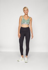 Sujetador deportivo verde claro con escote en V y tirantes anchos, combinado con leggings negros de cintura alta y zapatillas deportivas blancas.