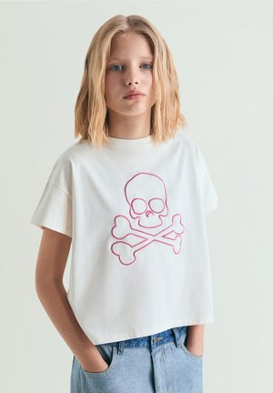 Chica rubia con camiseta corta blanca con diseño de calavera y huesos cruzados en rosa, de pie con las manos en los bolsillos de los vaqueros.