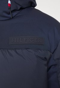 Marinblå puffjacka med en texturerad tyg, som har en gummibelagd "HILFIGER" logotyp patch på bröstet och dragsko på huva.