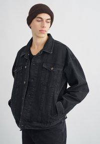 Casaco de ganga preto com corte descontraído, bolsos duplos no peito, botões prateados e acabamento ligeiramente desbotado. Usado com uma camisa escura e um gorro castanho.