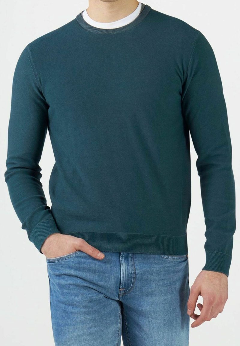 Mann trägt einen türkisfarbenen Pullover mit Rundhalsausschnitt und hellblaue Jeans, eine Hand in der Tasche, steht vor einem einfarbigen Hintergrund.