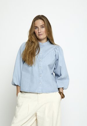 Žena s dlhými svetlohnedými vlasmi, oblečená v svetlomodrej blúzke s gombíkmi a nafúknutými rukávmi a krémových širokých nohaviciach na bielom pozadí.