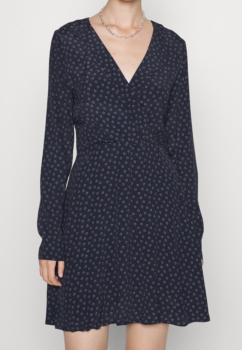 Femme portant une robe bleu marine à manches longues avec de petits motifs floraux clairs et un col en V, accessoirisée d'un collier chaîne en argent.