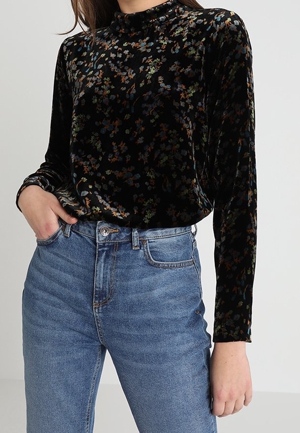 Kvinde iført en langærmet top i sort velour med et farverigt blomsterdesign, stoppet ned i højtaljede blå jeans.