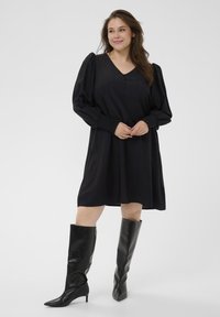 Sort kjole med V-hals, pufærmer og knapdetalje; parret med knæhøje sorte spidse støvler, der har en glat og skinnende tekstur.