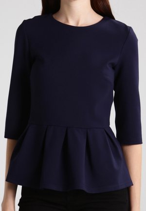 Vrouw draagt een donkerblauw marineblauw peplumtop met lange mouwen, een ronde halslijn en geplooide taille detail.