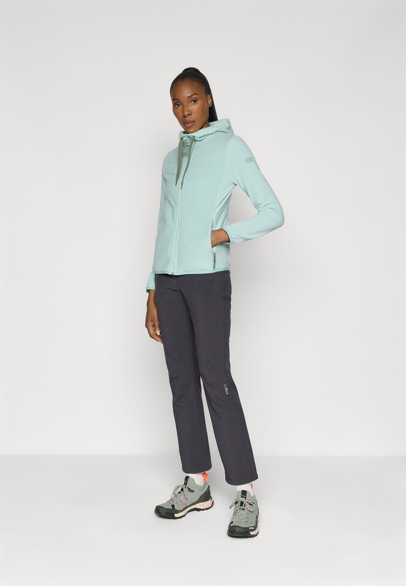 Helles blaues Fleecejacke mit Reißverschluss und Kapuze, strukturiertem Stoff; kombiniert mit dunkelgrauen Hosen und grauen Wanderschuhen mit pinken Akzenten.