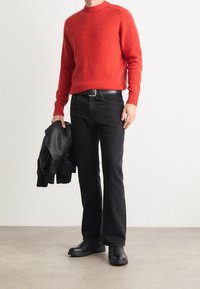 Roter Strickpullover mit Rundhalsausschnitt, schwarze Jeans und schwarze Stiefel. Eine schwarze Lederjacke ist über einen Arm gelegt.