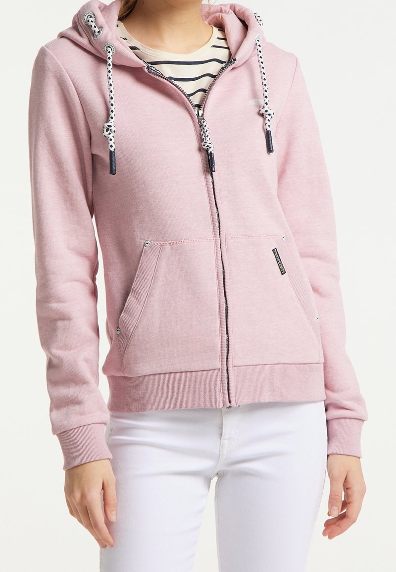 Femme portant un sweat à capuche rose clair zippé avec des cordons blancs sur une chemise rayée et un pantalon blanc, mains détendues le long du corps.