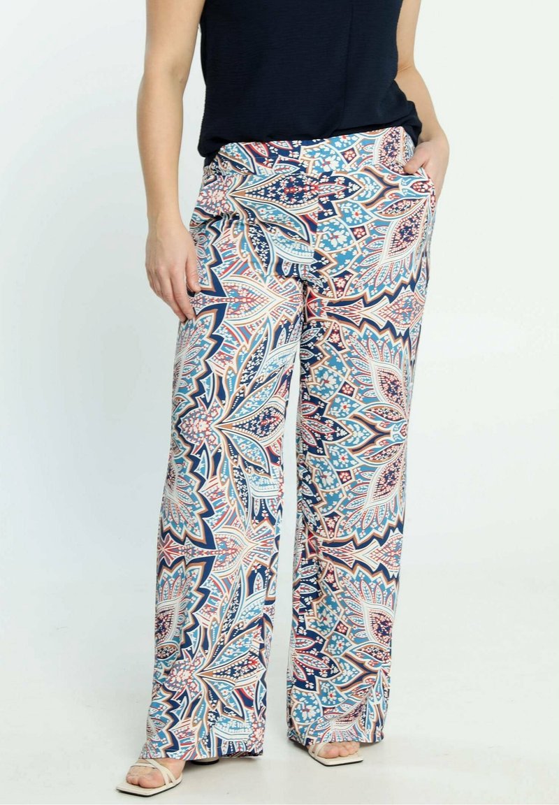 Femme portant un pantalon à jambes larges avec des motifs géométriques floraux bleus, blancs et roses, assorti à un haut noir et des sandales blanches.