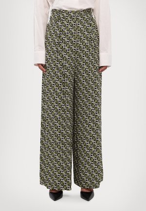 Pantalones de corte recto con un patrón geométrico en negro, blanco y verde, combinados con una camisa blanca de manga larga y zapatos negros puntiagudos.