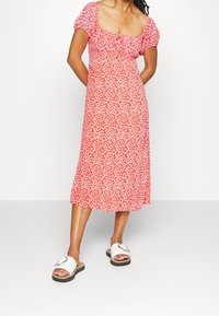 Robe fleurie rouge avec manches courtes bouffantes, corsage ajusté et jupe mi-longue. Portée avec des sandales blanches à enfiler ornées d'un accent métallique.