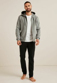 Grauer Reißverschluss-Hoodie mit Kordeln, über einem weißen T-Shirt getragen, kombiniert mit schwarzen Hosen. Die Textur wirkt weich und lässig. Barfußhaltung.