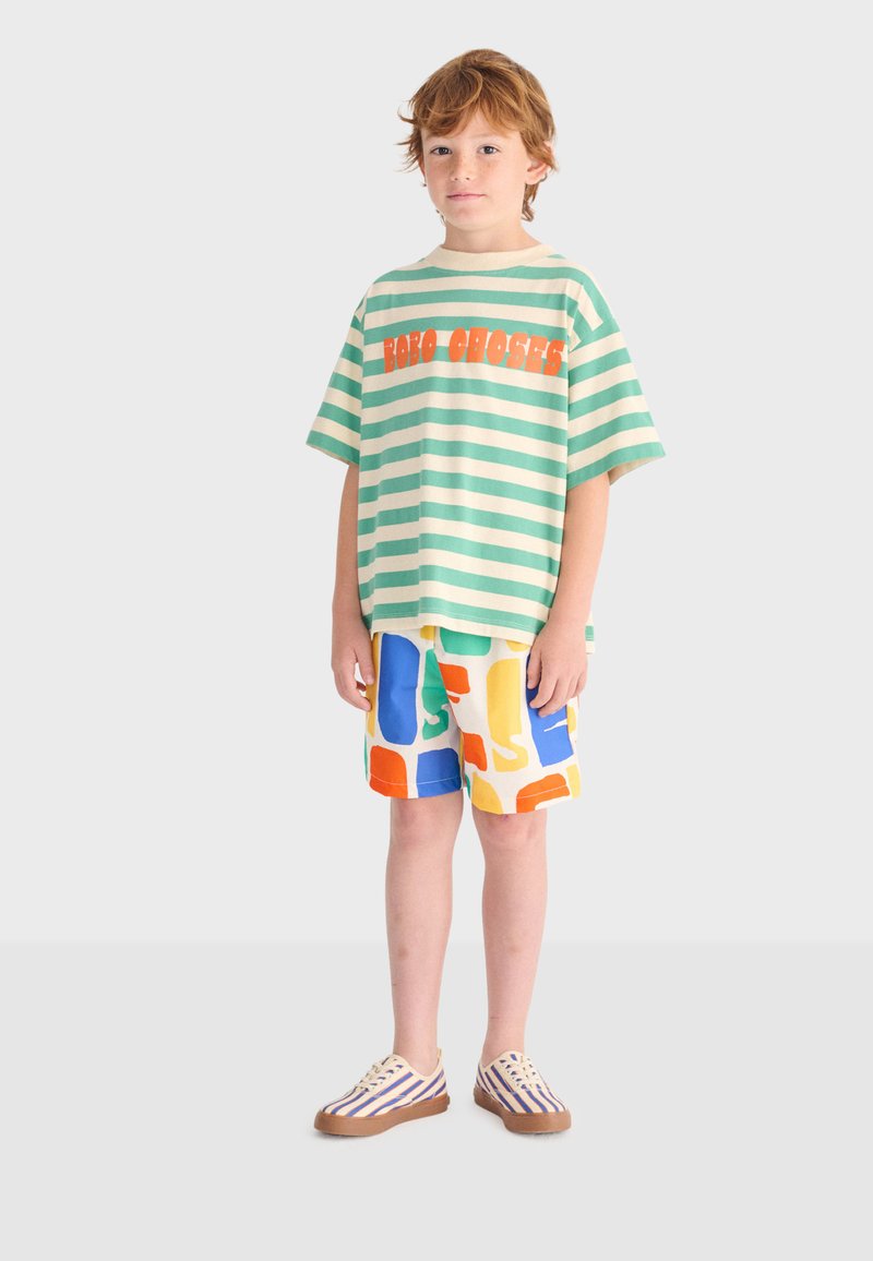 Junger Junge steht, trägt ein grünes und weiß gestreiftes übergroßes T-Shirt mit orangefarbenem Text, bunte gemusterte Shorts und gestreifte Turnschuhe.