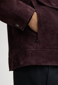 Giacca in suede borgogna con texture liscia, dotata di tasca laterale e dettaglio con bottone sul polso, abbinata a pantaloni scuri.