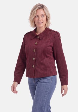 Vrouw met kort blond haar die een dichtgeknoopte bordeauxrode jas en blauwe spijkerbroek draagt, kijkt iets opzij met een ontspannen uitdrukking.